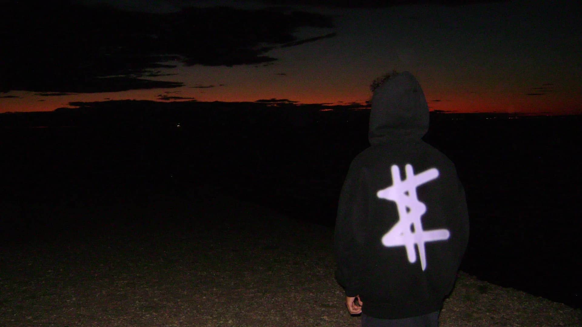 The EXSTSY glyph across the back of the black varsity zip hoodie, shot at last light